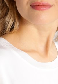 Weißes Baumwolloberteil mit weitem Ausschnitt und geripptem Kragen, präsentiert auf den Schultern eines Models, das eine glatte Hauttextur zeigt.