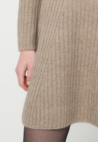 Bezs kötött ruha függőleges bordázással, hosszú ujjakkal, lekerekített szegéllyel és lágy tapintású; fekete harisnyával párosítva, a kéz a szegély közelében nyugszik.