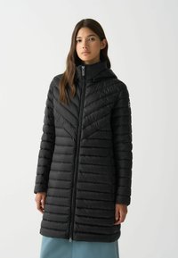 Melna pūkainā jaka ar ševronu rakstu, ar kapuci un priekšējo rāvējslēdzēju. Iezīmējas ar quilted sekcijām un gludu, spīdīgu tekstūru.