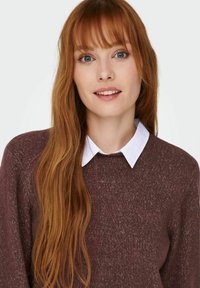 Mörkröd stickad tröja med långa ärmar och rundad hals, över en vit skjorta med krage. Långt, rakt, ljust brunt hår.