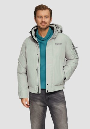 Donsjack in lichtgrijs, met een afneembare capuchon, zwarte accenten, ritszakken en een logo op de borst. Draag het over een teal hoodie.