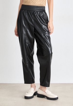 Pantalons en cuir noir avec une taille élastique, présentant une coupe décontractée et des détails de panneaux cousus. Portés avec des mocassins beiges.