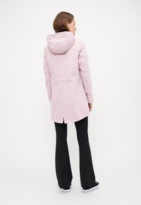 Lys rosa vanntett jakke med hette, med innsnørt midje, bakventil og rett silhuett, båret over svarte vide bukser.