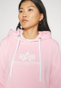Hoodie rosa realizzato in morbido tessuto, caratterizzato da un logo e testi bianchi "ALPHA INDUSTRIES" sul davanti. Include un cappuccio con cordini e una tasca a marsupio.