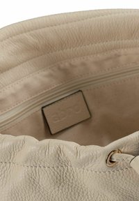 Interno di borsa a secchiello in pelle beige con tasca a cerniera e etichetta. Superficie texturizzata con apertura superiore arricciata e occhielli in metallo.