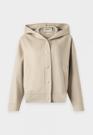 Personne aux longs cheveux foncés porte une veste beige boutonnée avec des manches larges et un pantalon ample vert olive clair sur un fond clair uni.