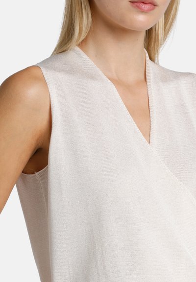 Moteris dėvi rankovių neturintį šviesiai smėlio spalvos megztą viršutinį drabužį su V formos iškirpte, parodyta nuo peties iki krūtinės vidurio priešais vienspalvį foną.