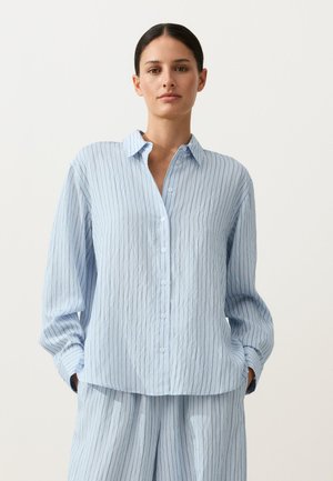 Femme portant une chemise boutonnée bleu clair et un pantalon ample assorti à fines rayures verticales, mains dans les poches, fond neutre.