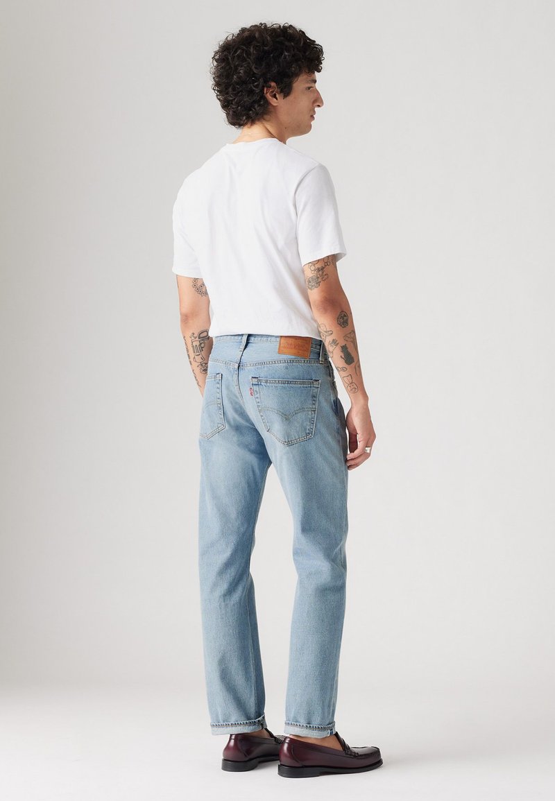 Hombre con cabello rizado y tatuajes vestido con una camiseta blanca, jeans de color azul claro y mocasines marrón oscuro, de perfil frente a un fondo simple.