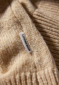 Tessuto di maglione beige chiaro, con una trama morbida, caratterizzato da un'etichetta bianca con la scritta "LINDBERGH" in testo nero.