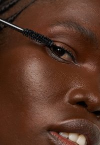 Du mascara noir est appliqué sur les cils d'un modèle à l'aide d'un pinceau noir. La peau est lisse, avec un éclat naturel et un maquillage subtil. Un œil marron clair est visible.
