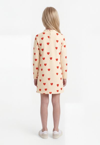 Fată văzută de la spate, purtând o rochie bej cu mâneci lungi și model cu inimioare roșii și adidași albi pe un fundal simplu.