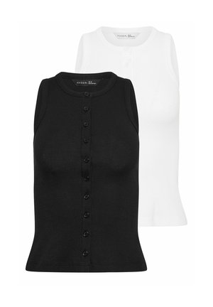 Femme debout, les mains dans les poches, portant un top noir sans manches boutonné, un jean bleu large avec revers et des baskets blanches.