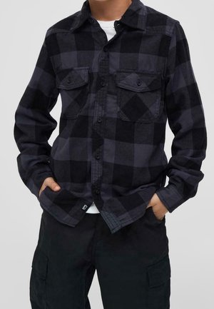 Person trägt ein schwarz-grau kariertes Hemd über einem weißen Unterhemd und schwarze Cargo-Hosen, die Hände in den Taschen.