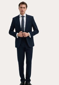 Marineblauw pak met een getailleerde pasvorm, button-up jasje, witte overhemden en een gestreepte stropdas. Zwarte schoenen maken de outfit compleet.