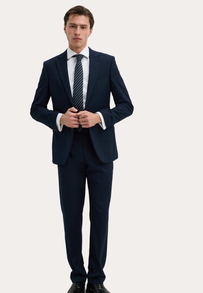 Marineblauw pak met een getailleerde pasvorm, button-up jasje, witte overhemden en een gestreepte stropdas. Zwarte schoenen maken de outfit compleet.