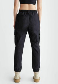 Pantaloni cargo neri con vita e polsini elasticizzati, dotati di tasche laterali e un tessuto texturizzato. Abbinati a stivali beige.