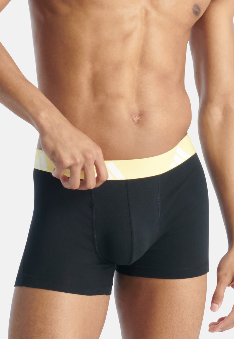 Boxer briefs negros con una cinturilla amarilla que presenta rayas diagonales blancas. La tela parece suave con una forma ajustada y costuras mínimas.