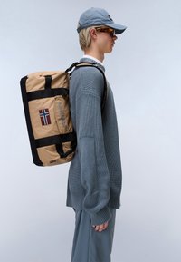 Un sac à dos beige et noir avec un patch de drapeau distinct, doté de bretelles robustes, d'un design à rabat et d'un tissu texturé et durable.