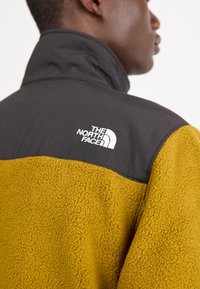 Giacca in fleece con parte superiore nera e parte inferiore senape. Include il logo The North Face in bianco sulla spalla. Tessuto a trama.