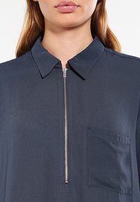 Blouse bleu marine avec fermeture éclair à l'avant, col pointu et poche poitrine unique. Texture douce et lisse avec une coupe décontractée.