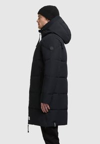 Abrigo acolchado negro con capucha, con secciones acolchadas, cremallera frontal y un parche de logo en la manga. Combinado con un gorro de punto negro.