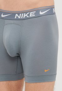 Pelēki bokseršorti, kas izgatavoti no gludas auduma, ar baltu Nike logo uz jostas un mazu oranžu logo apakšējā daļā.