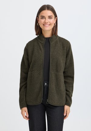 Jeune femme souriante, portant des boucles d'oreilles créoles dorées, une veste polaire vert foncé sur un t-shirt noir et un pantalon noir sur un fond clair uni.