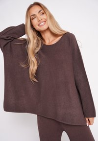 Brązowy, oversize sweter z dzianiny z długimi rękawami, szerokim dekoltem i miękką fakturą, noszony z pasującymi brązowymi dołem.