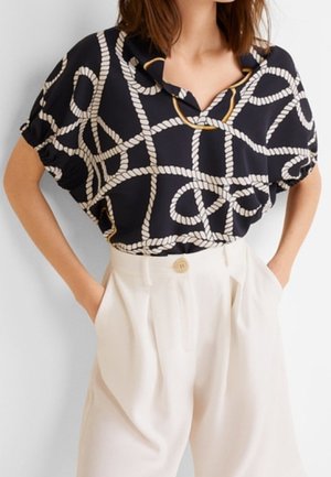 Blusa nera con motivo a corda bianca e accenti dorati; design largo con colletto. Abbinata a pantaloni crema a vita alta con un bottone.