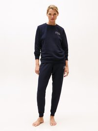 Marinblå sweatshirt och matchande joggers i mjukt tyg. Har ribbade muddar och midja, med en logotyp på bröstet. Barfota ställning.