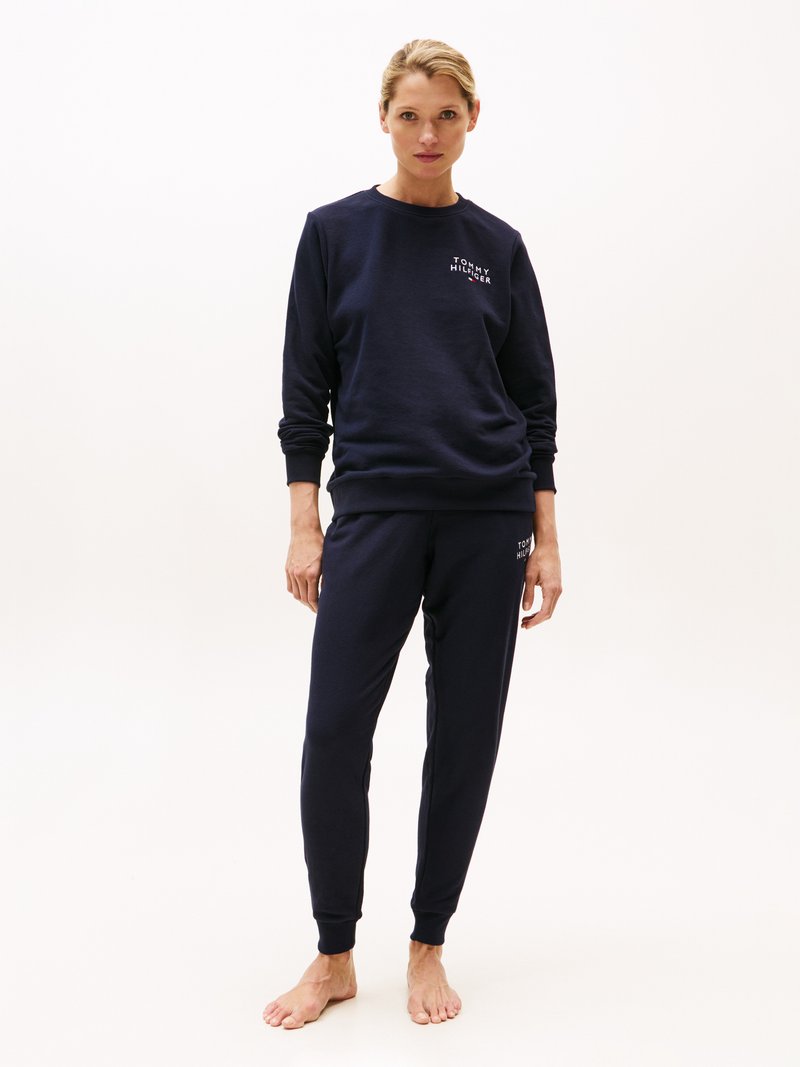 Marinblå sweatshirt och matchande joggers i mjukt tyg. Har ribbade muddar och midja, med en logotyp på bröstet. Barfota ställning.