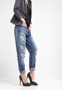 Osoba w podwiniętych, postrzępionych niebieskich dżinsach, czarnych szpilkach, czarnym topie i ciemnoszarym żakiecie, stojąca na tle białego tła.