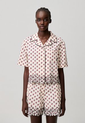 Mujer que lleva una camisa de manga corta color crema abotonada y pantalones cortos a juego con un pequeño estampado floral rojo y ribete de flor delineado en negro.