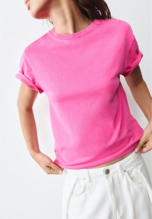 Kvinde iført en knaldrød kortærmet t-shirt med opsmøgede ærmer og hvide bukser, justerer taljebåndet.