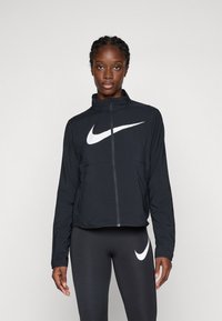 Frau steht da und trägt eine schwarze Nike Zip-Jacke mit weißem Swoosh-Logo sowie dazu passende schwarze Leggings mit weißem Swoosh-Logo.