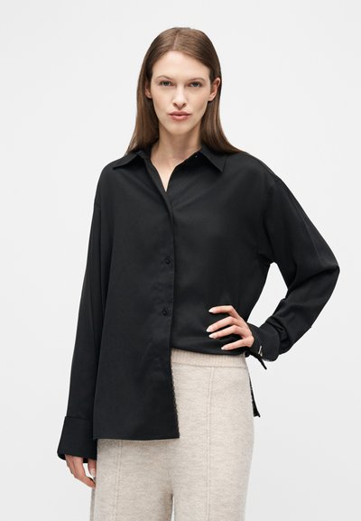 Γυναίκα με ίσια καστανά μαλλιά φοράει φαρδιά μαύρη πουκαμίσα με κουμπιά και μπεζ πλεκτό παντελόνι, στέκεται με το ένα χέρι στη μέση σε απλό φόντο.