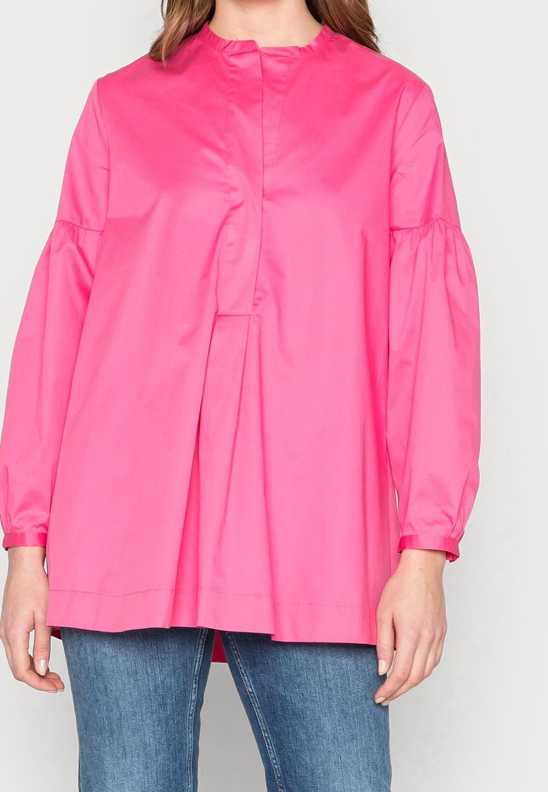 Femme portant un chemisier ample à manches longues de couleur rose vif avec un col haut et un jean bleu, shown du cou aux hanches.