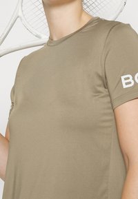 Camiseta deportiva verde oliva de manga corta, textura suave, escote redondeado y detalle de logo blanco en la manga izquierda. Raqueta de tenis de fondo.