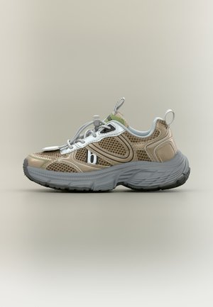 Baskets beige et blanches avec panneaux en mesh, semelle épaisse grise, languettes au talon et à la languette, et fermeture à lacets avec bouton-pression sur fond neutre.