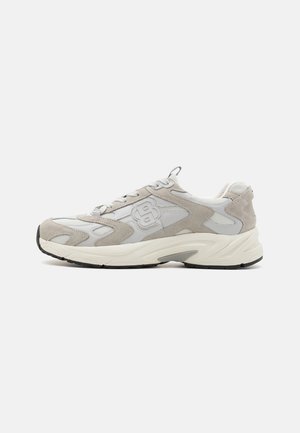 Sneaker atletica in mesh e suede grigio e bianco con suola chunky e chiusura con lacci, vista di lato su uno sfondo bianco.