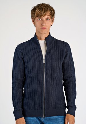 Marineblauwe ritscardigan met een gestructureerd kabelbreipatroon, hoge kraag, ribgebreide manchetten en zoom, gecombineerd met een eenvoudig wit T-shirt eronder.
