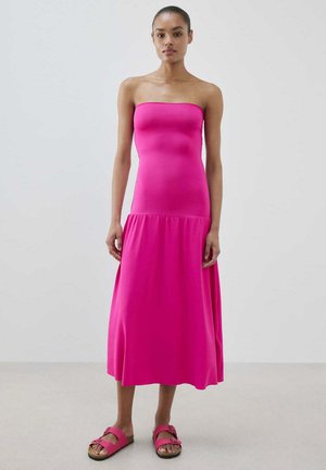 Žena oblečená v jasnoružových midi šatách bez ramienok a zodpovedajúcich ružových sandáloch stojaca na jednoduchej svetlosivej stene.