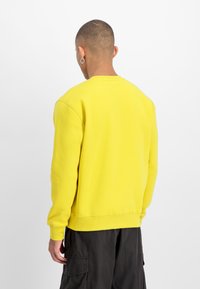 Gul genser laget av bomullsblanding, med lange ermer, ribbestrikkede mansjetter og en avslappet passform. Ingen synlige mønstre eller grafikk.