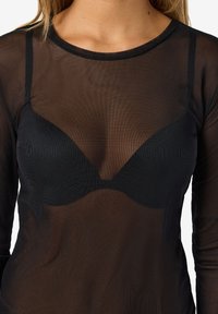 Ženska, ki nosi oprijet črn prosojen dolg rokav čez črni nedrček, prikazuje teksturo tkanine in podrobnosti plastenja.