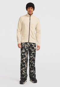 Lys beige fleecejakke med lynlås og sorte mønstrede bukser med creme detaljer. Model bærer en sort hue og sorte sko.