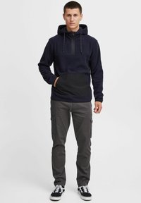Marineblauwe hoodie met een halve rits, zwarte voorzak en een getextureerd paneel. Gecombineerd met grijze cargobroek en zwarte sneakers.