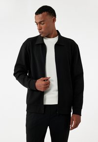 Sort zip-up jakke med kravedesign, blød tekstur og ribbede manchetter. Båret over en lysfarvet skjorte, der viser en afslappet pasform.