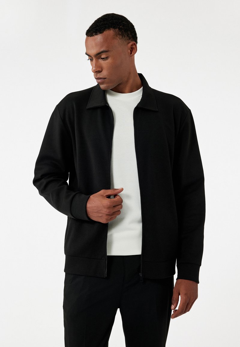 Sort zip-up jakke med kravedesign, blød tekstur og ribbede manchetter. Båret over en lysfarvet skjorte, der viser en afslappet pasform.