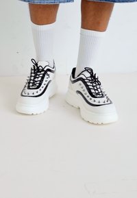 Zapatillas gruesas con un cuerpo blanco, detalles en negro y tachuelas metálicas. Incluyen cordones y suelas gruesas para mayor altura.
